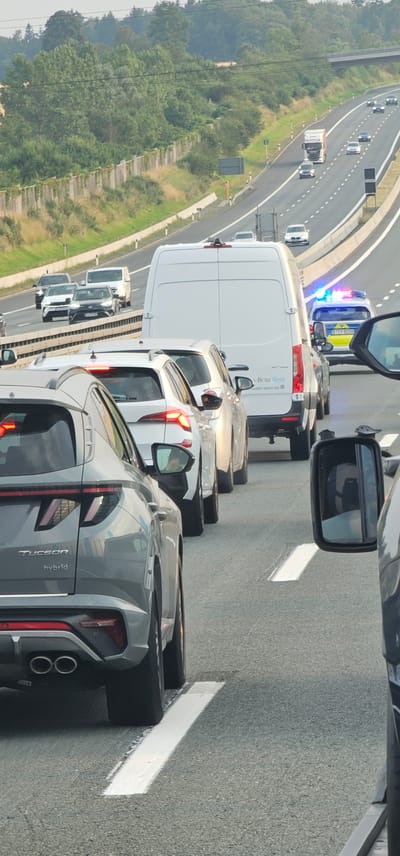The image captures a traffic jam on a multi-lane highway. In the foreground, a grey Hyundai Tucson Hybrid is visible from the rear, stopped in traffic. To its right, the rear of a white SUV with illuminated taillights is also stopped. Further ahead and to the right, a white Mercedes-Benz Sprinter van is parked, with a police vehicle displaying flashing blue lights immediately behind it. The police vehicle has license plate "BTZP 8058". The highway is lined with trees and vegetation on both sides, with a sound barrier visible to the left. A bridge crosses the highway in the distance. The scene is consistent with traffic congestion on a German autobahn.