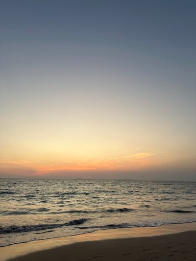 The scene displays a sunset view across a body of water from a sandy beach at Phra Ae, Thailand. The sky presents a vertical gradient, starting with deep blue at the top, transitioning to lighter blue, then pale yellow, and concluding with a horizontal band of orange and pink at the horizon. Several wispy clouds are visible within the orange-pink portion of the sky. The sea surface is dark and moderately textured with small waves, reflecting the warm colors of the sky. A strip of wet, light brown sand occupies the immediate foreground, showing reflections of the sky and faint human footprints. On the distant horizon, a landmass or island chain is visible as a hazy, dark silhouette towards the right. A single large, dark vessel or ship is discernible on the far left horizon. No individuals or explicit actions are present within the frame.