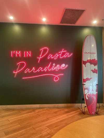 The image depicts an indoor scene with a dark green wall as the primary background. A neon sign on the wall displays the text "I'M IN Pasta Paradise" in bright pink. To the right of the sign, a surfboard stands upright on a black metal stand. The surfboard has a light-colored surface with a design of red and white clouds and waves. The text "VAPIANO" is visible in red at the top of the surfboard. The floor is made of light-colored wooden planks. The ceiling, illuminated by pink and white lights, has a rectangular vent. The setting does not specifically indicate the city of Surfers Paradise, Australia.