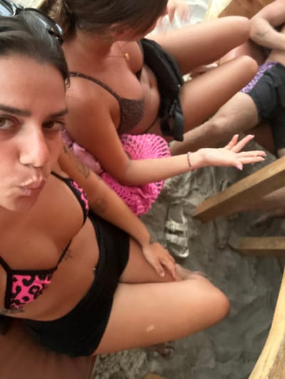 The image shows three people sitting on wooden chairs, positioned close to each other. Two women are wearing bikinis. The woman in the foreground is looking directly at the camera, smiling, and wearing a black skirt and a pink and black animal print bikini top. She is sitting on a wooden chair with her left leg bent and her right leg stretched out. The other woman is wearing a brown bikini top with a pink net top. She is sitting on a chair to the left of the foreground woman, and her right leg is resting on top of a man's leg, who is sitting on a chair behind her. The man is wearing black shorts with a white and blue design. They are on a sandy beach with a wooden table in the background. The scene is likely in Montañita, Ecuador, as the provided coordinates (latitude: -1.821, longitude: -80.756) correspond to a beach area in the town. The overall impression is one of casual relaxation and enjoyment.