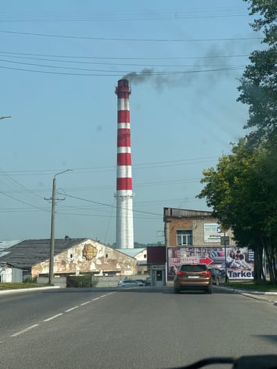 The image captures a street scene in Leninsk-Kuznetsky, Russia, on a clear, sunny day. A tall, red and white striped smokestack dominates the left side of the frame, emitting dark smoke. In the mid-ground, a brown Kia Sportage is parked on the right side of the road, facing away from the viewer, with its license plate reading "C280EP 142". To the right of the car, a brick building with a large advertisement featuring a family on a couch and the word "Tarket" is visible. Several other buildings and trees line the street, and power lines crisscross the sky. The road itself is asphalt with white dashed lines indicating lanes. The overall impression is of an industrial or semi-industrial area within the city.