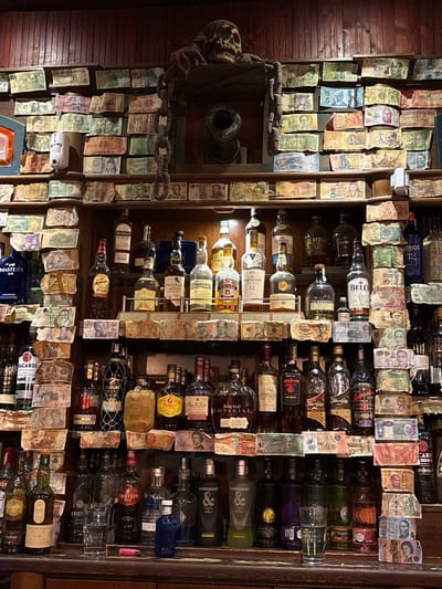 The image provides a detailed, eye-level view of the interior of a bar, specifically focusing on the shelves behind the counter. This location is identified as Taberna Casa Manteca in Cádiz, Spain, a establishment known for its distinctive decor.

The scene is dominated by a vast collection of international banknotes affixed to the dark wooden walls and shelving units. These currency notes, varying in size, color, and country of origin, are densely packed, forming a mosaic-like backdrop for the displayed beverages.

Multiple tiers of dark wooden shelves hold numerous bottles of various alcoholic spirits. Visible brands include Macallan, Talisker, Chivas Regal, Bell's, Aberfeldy, Master's Gin, Bacardi, Barceló Imperial, Gin Puerto de Indias, and Brugal. The bottles are arranged in an organized manner, with labels facing outwards.

On the top shelf, centrally positioned, a dark, stylized pirate skull wearing a red bandana rests. Below the skull, within a recessed alcove, a dark miniature cannon barrel is visible. Thick, dark, decorative chains hang from the skull's perch, extending downwards. The dark wooden bar counter in the foreground holds a few clear drinking glasses. No individuals, specific actions, or interactions are present in the image. The lighting suggests an indoor setting, likely provided by artificial sources, illuminating the display.