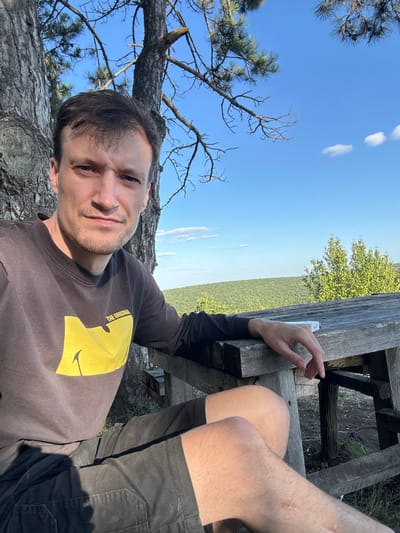 A man is taking a selfie while sitting at a wooden picnic table outdoors. He is wearing a dark long-sleeved shirt with a yellow graphic and black shorts. His right arm is resting on the table. The background shows a dense green forest under a clear blue sky with a few scattered clouds. The setting appears to be Velykozalissia, Ukraine, likely a park or natural area with picnic facilities.
