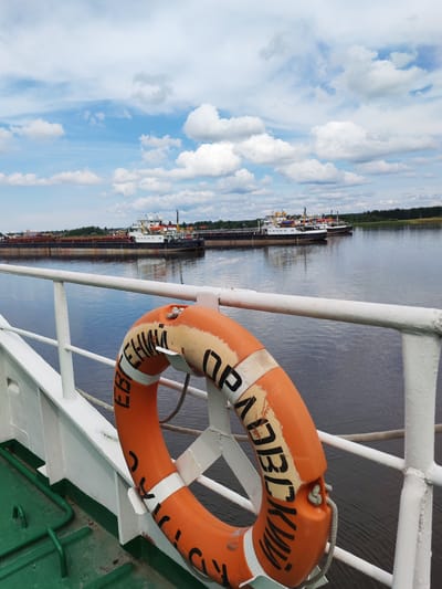 The image shows a view from the deck of a ship in the city of Shchetinino, Russia. In the foreground, an orange life preserver hangs on a white railing. Beyond the railing is a large body of water, with several barges and a cargo ship visible in the distance. The sky is blue with scattered white clouds. The environment appears to be a river or lake on a clear day. There are no people visible in the image. The scene is static and depicts a serene view of waterborne transportation.