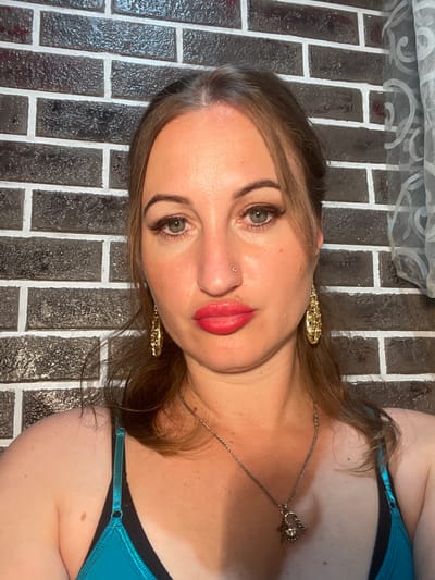 A woman with light brown hair and blue eyes is shown in a close-up selfie. She is wearing bright pink lipstick, gold earrings, and a silver necklace with a heart-shaped pendant. She is also wearing a teal-colored top. The background is a dark brick wall and a light-colored curtain with a swirling pattern. The image appears to have been taken indoors.