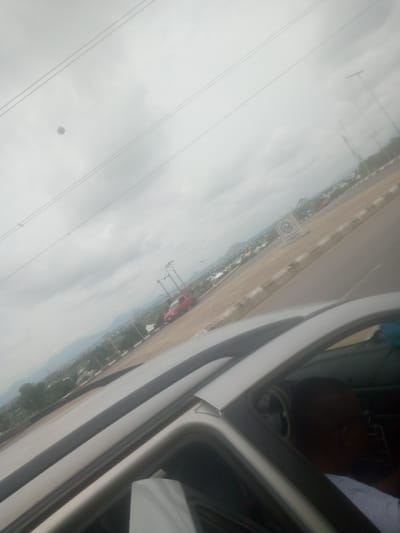The image captures a view from inside a vehicle, looking out at a road and a cityscape under a cloudy sky. The perspective is angled, with power lines stretching across the upper portion of the frame. In the distance, a town with buildings and trees is visible, with mountains forming a backdrop. A red car is parked on the side of the road. There is a sign with an emblem and text that appears to say "CRINBURG." The interior of the vehicle shows a person's head and shoulder, facing away from the camera. The overall scene suggests an urban or semi-urban environment in Lankoviri, Nigeria, with overcast weather.