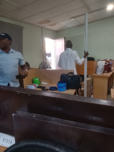 In Palmgrove, Nigeria, an indoor scene captures two individuals engaged in activity within what appears to be an office or workspace. On the left, a man wearing a blue collared shirt and a dark baseball cap is partially visible, facing away from the camera. To the right, another man in a white collared shirt stands facing away, holding a tall, white rectangular object, possibly a piece of construction material or equipment. The environment features multiple wooden desks and partitions, suggesting a cubicle setup. On one desk, a green bucket, a black bag, and a blue bucket are present, along with various small objects like containers and coiled cords. In the background, a fan and stacks of boxes are visible, and the room is lit by overhead lighting. The overall impression is one of ongoing work or setup within a functional, somewhat cluttered, indoor space.