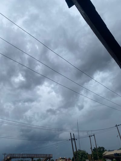 The image displays a heavily overcast sky dominated by dense, dark grey clouds, suggesting stormy or impending rainy weather. Multiple utility lines, likely power or telephone lines, stretch diagonally across the frame from the middle-left towards the upper-right.
On the left side of the image, several wooden utility poles are visible, supporting these overhead lines. Behind the poles, glimpses of green foliage from trees or bushes, and parts of buildings or structures (including what appears to be scaffolding or a building facade), can be seen, indicating an urban or semi-urban environment. A dark, angled structure, possibly a roof edge or a wooden beam, occupies the bottom-right corner of the frame. Small, dark specks, potentially birds or insects, are discernible against the brighter areas of the clouds.
The scene conveys a somber, dim daytime atmosphere under a thick cloud cover. The weather is the prominent feature, hinting at a recent downpour or an impending storm. The visible infrastructure—utility poles and lines—situates the scene in a populated area. This imagery reflects a cloudy day in Auchi, Nigeria. There is no discernible text within the image.