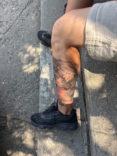 This image provides an overhead view of a person's lower body in an outdoor setting within the city of Tenevo, Bulgaria. The subject's bare legs display a tanned skin tone. The left lower leg is adorned with a large, detailed black and grey tattoo portraying a classical-style face or figure, featuring warm reddish undertones, likely from illumination or the tattoo's pigment. The person wears light-colored, likely linen, shorts visible on the upper right thigh and black New Balance 1000 series athletic shoes on both feet. These shoes are identifiable by the "1000" marking on the lateral midsole and a chunky sole design, constructed with mesh and synthetic materials. The scene is under direct sunlight, producing clear, dappled shadows on the ground and legs, consistent with overhead foliage. The ground surface is composed of concrete paving, showing variations in texture, cracks, and shades. A discarded cigarette butt is visible on the concrete near the left shoe, alongside small fragments of dried vegetation in the pavement cracks.