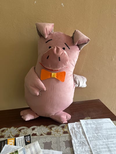 A plush toy pig, pink with a simple face and an orange bow tie, sits on a wooden surface. The pig is positioned centrally and faces forward, its ears tilted slightly to the sides. To the right of the pig, a portion of its arm is visible. The wooden surface is cluttered with several white documents, some partially obscured by the pig and a decorative lace runner. The background consists of a textured, pale yellow wall. The overall scene appears to be indoors, possibly in a room within the city of Gavrilov Posad, Russia, given the context of the image.