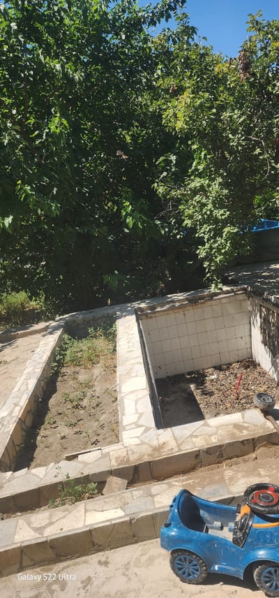 The image depicts an outdoor scene in the city of Vabkent, Uzbekistan, characterized by a partially constructed or neglected outdoor area. In the foreground, a blue toy car is parked on a concrete surface. Beyond the car, there are tiered concrete structures, some of which are lined with irregular stone tiles. Two rectangular depressions are visible within these structures. One depression contains sparse, young green plants growing in dry soil. The other depression appears to be a tiled pit filled with dried leaves, debris, a red stick, and a small black wheel. Lush green foliage from trees overhangs the scene from above, and the bright blue sky is visible through the leaves. The overall impression is one of an unfinished or overgrown outdoor space.