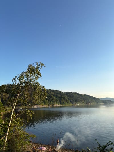 The image depicts a serene lakeside scene in Lipie, Poland. In the foreground, a slender birch tree with delicate leaves leans towards the tranquil water. The shoreline is a mixture of sand and pebbles, where a few individuals are visible. One person stands near a small fire producing wisps of smoke, while another sits beside a pink umbrella. The lake stretches out, reflecting the clear blue sky and the lush green hills that form the backdrop. Several boats are anchored in the distance, adding to the peaceful atmosphere. A faint crescent moon is visible in the upper right corner of the sky.