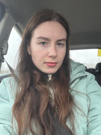 The image captures a fair-skinned female, an adult, situated in the front passenger seat of a vehicle in the city of Gurlevo, Russia. She possesses long, wavy light-to-medium brown hair with subtle auburn tones, light-colored eyes (blue-grey), and light pink lipstick. A small gold hoop earring is visible in her left ear. She is dressed in a light mint green or pale turquoise puffer jacket over a darker garment, with a beige zipper pull evident at the collar of an inner layer. A dark seatbelt crosses diagonally over her left shoulder and chest. The vehicle's interior features a grey headliner and a black headrest of the rear passenger seat. Through the vehicle's windows, a hazy, overcast outdoor environment is visible, displaying bare trees or foliage and indistinct distant structures. A rectangular yellow sticker is affixed to the upper right section of the rear window. The subject is looking directly at the camera.