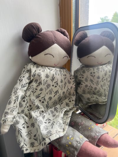 A stuffed doll with brown hair in two buns is sitting next to a mirror, appearing to look at its reflection. The doll is wearing a white dress with a black floral print and grey patterned pants. It has a cloth face with closed eyes and a small pink smile. A white bead earring is visible on its left ear. The doll is positioned on a wooden surface, likely a windowsill, with a white wall to its left and a window with a view of greenery to its right. The mirror, which is rectangular with rounded corners and a dark frame, reflects the doll's head and upper body. The scene is brightly lit, suggesting it is daytime. The overall impression is that of a whimsical and quiet setting.