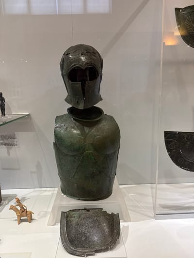 The image displays a museum exhibit in Heraklion, Greece, featuring ancient bronze armor and artifacts. The central subject is a damaged bronze helmet and cuirass, likely from a Greek warrior, positioned on a transparent stand. To the left of the armor, a small, terracotta figurine of a person riding a horse is visible on a lower shelf, next to informational text. In the background, through a glass display case, another section of the museum is visible with a fragment of bronze weaponry. The lighting is soft and diffused, typical of a museum setting, illuminating the aged patina of the bronze objects. The overall scene is one of historical preservation and display.