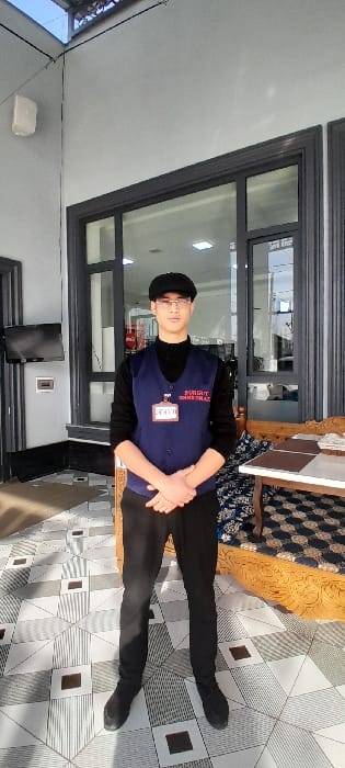 The image displays a young man standing indoors, appearing to be posing for a photograph. He is oriented vertically in the image, suggesting the photo might have been taken in portrait mode or rotated.
The man has a youthful appearance, wearing glasses and a black cap. He is dressed in a black long-sleeved shirt, over which he wears a dark blue vest. On the left side of his vest (viewer's right), white text is visible, partially reading "BUDGET OVERNIGHT" vertically. Below this, a red-bordered white name tag is attached, with the numbers "422" clearly visible, and possibly other, less distinct text. His hands are clasped together in front of him. He wears black pants and black shoes.
The setting appears to be an indoor space with a modern aesthetic. Behind the man, large windows with dark grey frames are visible, reflecting interior lights or the outdoor environment. To the man's right, a patterned wooden bench or low couch with a blue and white patterned cushion is visible, contributing to the room's decor. The floor features a striking geometric pattern of light grey, white, and darker grey squares and rectangles. In the background, to the man's left, a dark flat-screen television is mounted on a light-colored wall, and a white rectangular object, possibly a speaker or light fixture, is also attached to the wall.
The lighting in the room is bright, with shadows cast on the patterned floor, indicating a strong light source, possibly natural light from the windows combined with artificial interior lighting. No specific time of day or location is explicitly apparent, but the overall impression is of a clean, well-maintained commercial or institutional indoor space. The man's attire, particularly the vest with text and a name tag, suggests he might be an employee or volunteer.