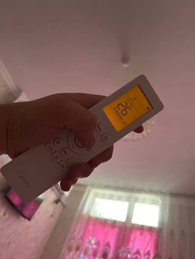 An over-the-shoulder shot captures a person's hand holding a white Midea brand air conditioner remote control. The remote's screen displays a set temperature of 26 degrees Celsius, with the "COOL" mode active and an "AUTO" fan speed indicated by vertical lines. Below the screen, various buttons are visible, including "Mode," "Turbo," "Swing," "LED," and "Clean," along with directional arrows. The remote is angled upwards, suggesting it's being used to control an appliance positioned higher up. The background is out of focus, showing a room with windows and curtains, illuminated by natural light. The image's lighting and the soft focus suggest an indoor environment. The city of Türkistan qalası, Kazakhstan is not depicted in this image.