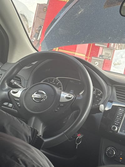The scene depicts the interior of a vehicle, viewed from a seated position, looking forward through the windshield. A prominent black Nissan steering wheel occupies the central foreground, featuring silver trim, control buttons (e.g., "CRUISE ON/OFF"), and a central Nissan logo. Behind the steering wheel, a portion of the instrument cluster is visible, displaying a digital time of "4:32" and a temperature reading of "33." A set of car keys with a remote fob hangs from the ignition or key slot to the right of the steering column. The leg of an individual, clad in dark trousers or fabric, is visible in the lower left foreground.
Through the windshield, a gas station environment is observed. Multiple red and white striped gas pumps are visible, with one pump clearly displaying the number "2." The ground in the exterior scene shows areas covered with snow or slush. A dark-colored vehicle hood, appearing dusty, is partially visible in the upper central portion of the windshield view. A brown building or structure is partially visible behind the gas pumps on the left. This scene is situated in the city of Apnagyugh, Armenia.