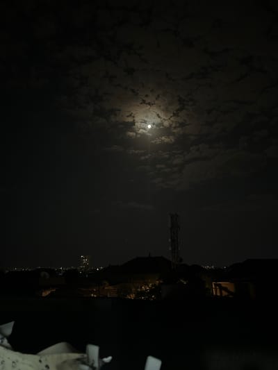 The image depicts a night scene in the city of Sabunchu, Azerbaijan. A bright full moon is visible in the sky, partially obscured by scattered clouds. Below the moon, a tall communication tower stands prominently. In the middle ground, there are the dark silhouettes of buildings and rooftops, with some distant city lights and a lit-up high-rise building visible on the horizon. The foreground is dark and indistinct, with the suggestion of some objects or structures. The overall atmosphere is one of a calm, moonlit night over a residential or semi-urban area.