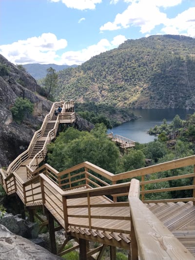 A scenic overlook point in Alijó, Portugal, presents a wooden staircase winding down a rocky hillside toward a body of water. The steps have wooden railings and are staggered to adapt to the terrain. A couple of people are walking on a section of the stairs further up the hill. The background reveals a mountain covered in trees and a river or lake. The sky is blue with scattered clouds.