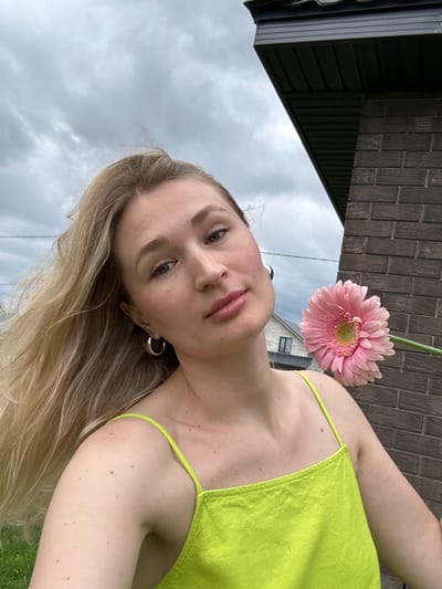 A close-up, selfie-style photograph shows a young woman with long, blonde hair and light green eyes. She is wearing a bright green, spaghetti-strap top. A large, pink gerbera daisy is held near her face, with its stem extending out of frame. The woman is positioned outdoors, with a brick wall to her right and a cloudy sky in the background. In the distance, a house with a dark roof is visible. The woman is looking directly at the camera, with a neutral expression. The image appears to have been taken in the city of Mokino, Russia.