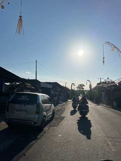 An outdoor, eye-level, full shot captures a street scene in Kubu, Indonesia, on a clear day with the sun high in the sky. The scene is dominated by a road stretching into the distance, flanked by buildings and overhead power lines. A silver sedan is parked on the left side of the road, its rear facing the camera, with a "PREDATOR" sticker on the back window and a license plate that reads "DK 4074 KW". Several people are riding motorcycles down the road, their forms silhouetted against the bright sun. The motorcycles are heading away from the camera. On either side of the road, arched structures with hanging decorations are visible, suggesting a festive or religious occasion. The sky is a brilliant blue with a few wispy clouds. The overall impression is one of a typical day in a bustling Indonesian town, bathed in the warm light of the sun.