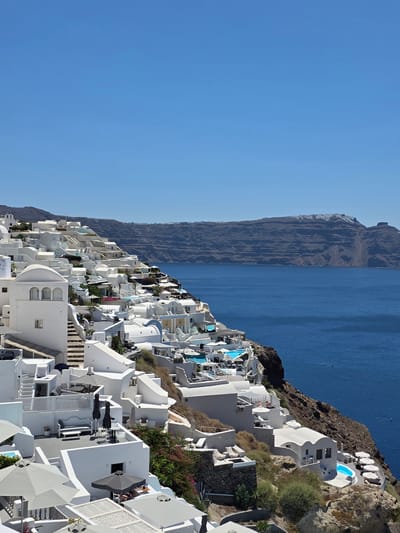 This media file captures a scenic view of a whitewashed village cascading down a steep cliff face overlooking the deep blue Aegean Sea. The architecture is characteristic of Greek island villages, with cubic buildings and rounded edges, adorned with vibrant blue-domed swimming pools. Some of these pools appear to be private, integrated into the terraces of the white structures.
The village is built on a dramatic rocky incline, with lush green vegetation and flowering bushes interspersed among the buildings, adding touches of color to the predominantly white and brown landscape. The sea is calm and vast, stretching out to meet a rugged, mountainous coastline in the distance, which shows signs of erosion and layered rock formations. The sky is a clear, brilliant blue, suggesting a sunny day, likely in the late morning or early afternoon given the bright lighting and strong shadows.
Scattered throughout the village are signs of leisure and relaxation: lounge chairs and umbrellas are visible near the pools and on various terraces. A few people can be seen enjoying the ambiance, suggesting a popular tourist destination. The overall atmosphere is one of tranquility and picturesque beauty, embodying a quintessential Greek island experience. There is no visible text in the media file.