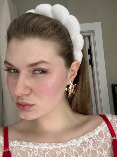 A fair-skinned person wearing a white, lace top with red straps is angled in a portrait. The person has light brown hair partially covered by a white, cloud-like headband. They wear a star-shaped earring adorned with multiple colored jewels. Their skin has a subtle blush, and their lips are glossy. The background shows a neutral-colored wall and a portion of a white door frame.