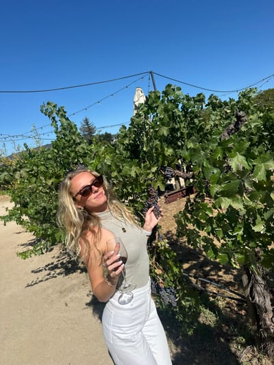 A young adult female is depicted standing outdoors in a vineyard, identified as being in Rutherford, United States. The scene is illuminated by bright sunlight under a clear blue sky. The subject, positioned slightly left of center, possesses long, wavy blonde hair and wears dark sunglasses, a light-colored sleeveless mock-neck top, and white pants. She smiles directly at the camera. Her right hand holds a clear wine glass filled with red wine, while her left hand gently touches a cluster of dark grapes on an adjacent vine. A light-colored dirt path is visible in the foreground. Rows of vibrant green grapevines laden with dark grapes extend throughout the mid-ground and background. Overhead, string lights are suspended between poles. In the background, a large, light-colored umbrella or canopy structure is partially visible, along with a conifer tree and distant hills.