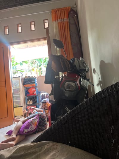 The image depicts an interior scene in what appears to be a residential setting in Serdang Kulon, Indonesia. A person is lying on the floor, propped up by a purple floral pillow, with their legs extended. They are wearing a dark floral dress and a blue garment on their upper body. To the left, a wooden door is open, revealing a glimpse of lush greenery and bright daylight outside. Inside the room, a multi-tiered blue shelf unit contains various items, including shoes and bags. A refrigerator is also visible to the left of the shelf. In the center of the image, a red and black motorcycle is parked. Clothes are draped over the handlebars of the motorcycle. A dark woven furniture piece occupies the foreground on the right. The walls are painted a light color, and orange curtains are visible near the open doorway. Small rectangular ventilation openings are present high on the wall.