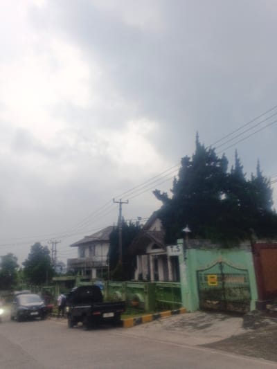 The image portrays an outdoor street scene in the city of Morrumbala, Mozambique, under an overcast sky. A paved road stretches across the lower portion of the frame. On the left side of the road, a dark-colored sedan or hatchback is stationary, exhibiting illuminated headlights. Adjacent and to the right of the sedan, a dark-colored pickup truck with an open bed is also stationary on the road's shoulder, facing away from the viewer. Two individuals, indistinct due to distance and resolution, are positioned between these vehicles, appearing to stand near the front of the pickup and the side of the sedan.

To the right of the road, a light green wall and a matching metal gate delineate a property. A white sign with the letters "T.S" is mounted above the gate. Behind this boundary, several tall, dark green coniferous trees are prominent. Further to the left along the property line, a building with a peaked roof and white columns at its entrance is visible. Behind this, a larger, two-story light-colored house featuring an upper-level balcony can be seen. Multiple utility poles supporting overhead wires are distributed across the midground. The environment is characterized by diffuse natural light consistent with heavy cloud cover.
