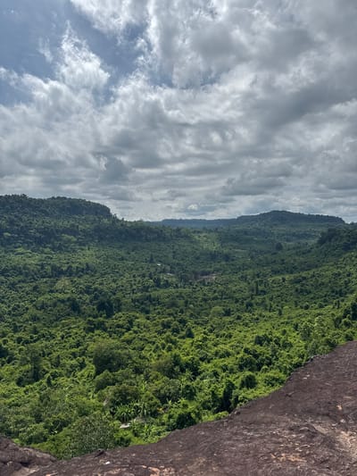 The image shows a lush, green, dense jungle covering rolling hills and valleys under a cloudy sky. In the foreground, a large, dark, rocky outcropping occupies the bottom right corner, with the texture of weathered stone visible. The jungle vegetation is varied, with different shades of green indicating diverse plant life, including trees, vines, and possibly some banana plants in the lower sections. The terrain appears rugged and natural, with no visible signs of human development or habitation within the jungle itself, except for what might be a faint, unpaved path or clearing visible in the mid-ground. The sky is filled with dramatic, gray and white clouds, suggesting an overcast or partly cloudy day, with patches of blue sky visible through the gaps. The overall impression is one of a remote, untouched natural landscape. The location is identified as Banteay Srei, Cambodia, a region known for its verdant tropical environment. There are no people or animals depicted in the image.