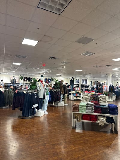 The image shows the interior of a clothing store. Mannequins dressed in various outfits stand in a line, positioned between rows of folded garments on shelves. The floor is made of wood planks. Overhead, a ceiling comprised of square tiles is visible, with various light fixtures and ventilation panels. The store appears well-lit and organized.