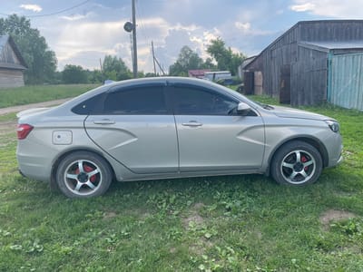 A light-colored sedan is parked on a grassy area next to a wooden barn and other rustic structures. The car has tinted windows and custom rims with red brake calipers. The sky is cloudy with patches of blue. The scene appears to be in a rural or semi-rural setting, possibly in Nizhnii Bugrysh, Russia.