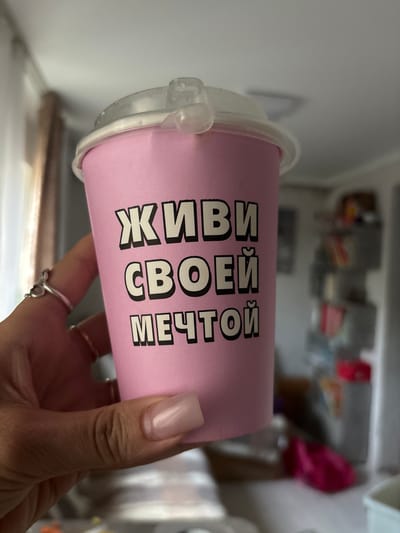 A close-up, vertically oriented shot features a pink disposable cup with a clear plastic lid, held by a person's hand. The cup has black, bold, block lettering printed vertically. The visible text reads "ЖИВИ СВОЕЙ МЕЧТОЙ". The cup appears to be inside a refrigerator, with shelves and various food items blurred in the background. The lighting is bright, suggesting the refrigerator light is on. The person's fingernails are long and painted a light, neutral color. They are wearing several rings on their fingers. The overall impression is a casual, everyday moment, perhaps during meal preparation or storage in a kitchen. The location context of Zaşağan, Kazakhstan does not provide any additional visual cues within the image itself.