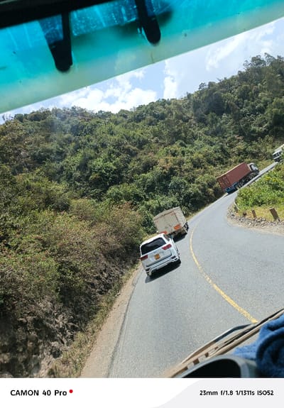 A winding asphalt road with a yellow dividing line curves through a lush, green hillside. A white SUV follows closely behind a box truck. A larger container truck is visible further up the road. The view is from inside a vehicle, evident from the interior elements at the top and bottom of the frame, including the rearview mirror and parts of the dashboard. The landscape features dense vegetation and rolling hills under a partly cloudy sky.