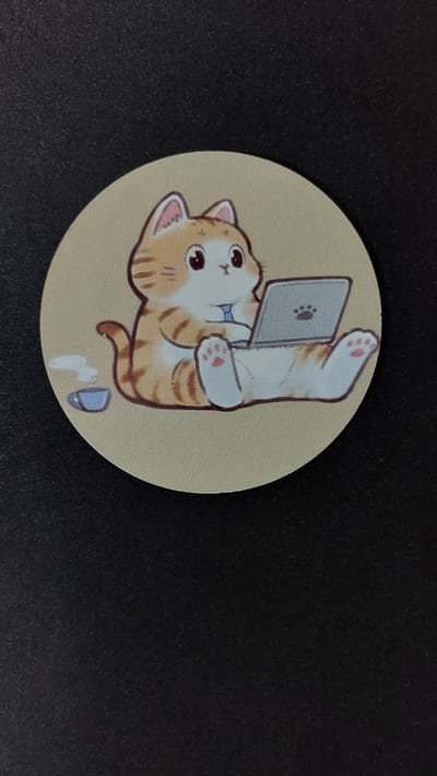 The image shows a circular sticker with a cartoon drawing of an orange and white tabby cat sitting on its hindquarters and using a laptop. The cat is wearing a blue bowtie. To the left of the cat is a small blue teacup with steam rising from it. The sticker is placed on a dark, textured surface, likely a desk or table. The background is black and out of focus. There is no indication of any specific location in Oleśnica, Poland, or any other city.