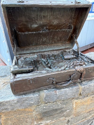A bronze sculpture of an open suitcase is mounted on a stone wall. The suitcase is detailed with a handle, hinges, and latch. The interior of the suitcase contains sculpted items including a bottle, possible clothing, and a camera. The open lid of the suitcase reveals a textured, empty interior. The stone wall provides a rough, natural contrast to the polished bronze.
