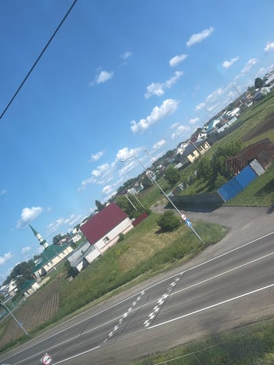The image shows a village scene in Kamaevo, Russia, under a bright blue sky with scattered white clouds. A road runs diagonally across the foreground, with white dashed lines indicating lanes and some speed bumps. To the left of the road, a grassy embankment slopes upwards, with several houses and a mosque with a green roof and minaret visible. Further back, more houses with various roof colors and agricultural fields can be seen. A tall street lamp stands near the road, and a blue metal fence borders some of the properties. A red-bricked building with a red roof is prominently featured on the right side of the image, with a portion of a corrugated metal structure and a blue metal fence nearby. The overall impression is of a rural, residential area on a sunny day.