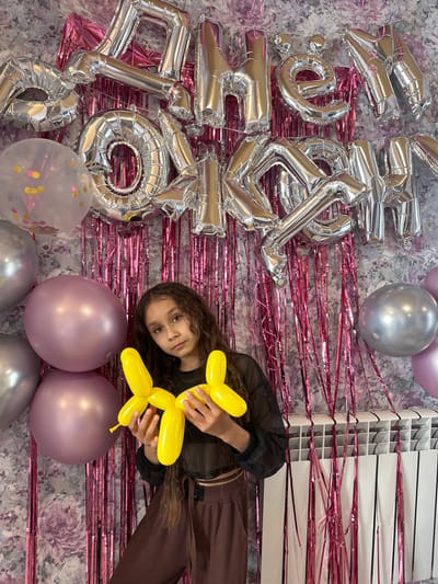 In a room with floral wallpaper, a young girl with long, dark, curly hair stands in front of a backdrop of silver foil balloons spelling out "С ДНЕМ РОЖДЕНИЯ" (Happy Birthday) and pink tinsel curtains. She wears a sheer black top and brown pants. In her hands, she holds a yellow balloon dog. To her left are several large, round balloons in shades of silver and mauve. To the right of the girl is a white radiator covered partially by the pink tinsel. The scene suggests a birthday celebration.