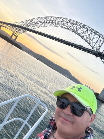 The image depicts a male individual taking a selfie from a boat, positioned in the lower right foreground. He is wearing a neon yellow baseball cap with a dark logo, dark-framed sunglasses, and a red, white, and green plaid shirt. A thin, dark necklace is visible. The individual has a slight smile. A white metal boat railing occupies the left foreground.
In the background, the Bridge of the Americas, an arched truss bridge, spans a wide body of water, likely the Panama Canal or Bay of Panama, within the Ancón district of Panama City, Panama. The sky behind the bridge exhibits a sunset or dusk coloration, with warm orange hues near the horizon transitioning to lighter tones above. Distant hills or a coastline are visible on both sides of the water, with what appear to be industrial cranes on the far bank to the right. The water's surface is rippled, reflecting the ambient light.