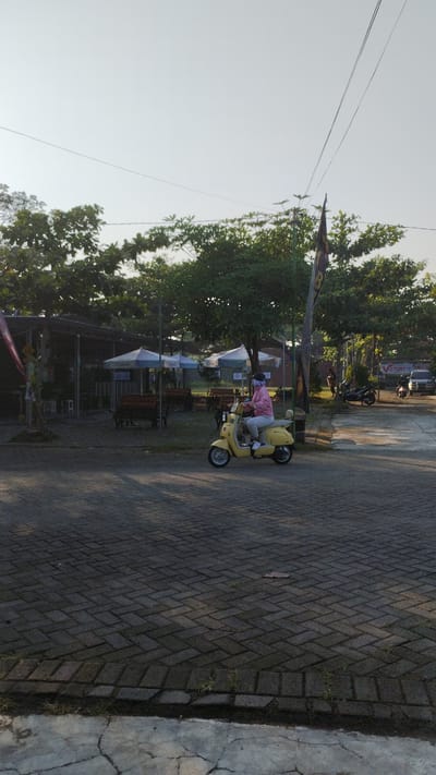 A medium-shot, eye-level, exterior, daylight, vertical, full shot, captured in Trosobo, Indonesia, shows a person riding a yellow scooter on a brick-paved area. The scooter rider, wearing a pink jacket and grey pants, has their face obscured by a light-colored mask and a dark fabric covering their head. The scooter has a black seat and a white rear storage compartment. In the background, there are several outdoor seating areas with tables and chairs under white umbrellas, suggesting a food or recreational establishment. Trees with green foliage surround the area, and power lines are visible crisscrossing the sky. A dark flag with yellow lettering is planted on a pole to the right of the scooter. Further in the background, on the right side of the image, a white SUV and a dark motorcycle are parked, with a sign that reads "VICTORY 15" visible on a building. The overall lighting suggests it is either early morning or late afternoon, with long shadows cast on the ground. The brick pavement is laid in a herringbone pattern.