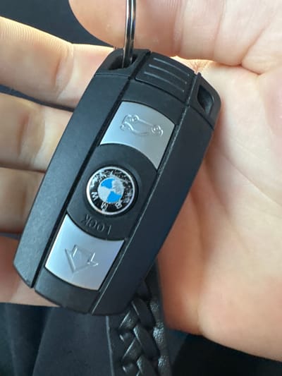 A hand holds a black BMW car key. The key features silver buttons with car, BMW logo, and lock icons. A black braided leather strap is partially visible. The background is out of focus. The location is not discernible from the image.