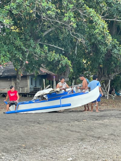 Four men are gathered around a white and blue outrigger canoe on a sandy beach in Dauin, Philippines. The canoe is positioned on the sand, angled towards the viewer's right.
The man on the far left, wearing a red t-shirt and patterned shorts, is seated on the edge of the canoe, facing left. He appears to be looking off to the side.
In the center of the image, a man with a patterned shirt is seated inside the canoe, facing left and holding what appears to be a musical instrument, possibly a ukulele. To his right, a bare-chested man is standing and looking at the canoe. Behind him, another man in a blue t-shirt and shorts stands, also looking towards the canoe.
Behind the men and the canoe, there is lush green foliage from large trees. In the background, to the left, a small, rustic structure with a thatched roof and shuttered windows is visible. The ground is covered in dark sand, with scattered small pebbles and debris. The overall scene suggests a relaxed, outdoor setting.