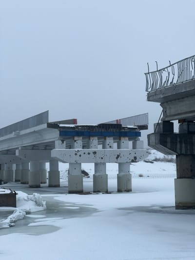 The image depicts a severely damaged multi-span concrete bridge spanning a snow-covered, partially frozen body of water in Staryi Saltiv, Ukraine, under an overcast sky. The bridge exhibits extensive structural failure, with a large central section entirely missing, creating a significant gap. The concrete bridge deck on the far right terminates abruptly mid-air, and its metal safety railing is severely twisted and bent upwards. The concrete support structure of the adjacent central bridge segment shows numerous pockmarks and impact damage. To the left, a section of the concrete bridge with an intact metal guardrail extends towards the void. Exposed rebar and internal structural elements are visible at the points of fracture. The terrain below and around the bridge is uniformly covered in a layer of snow and ice, with visible patches of dark open water. A dark, rectangular object rests partially submerged in the ice on the left foreground. Barbed wire is visible on the snow-covered embankment in the background to the right. No individuals are present in the scene.