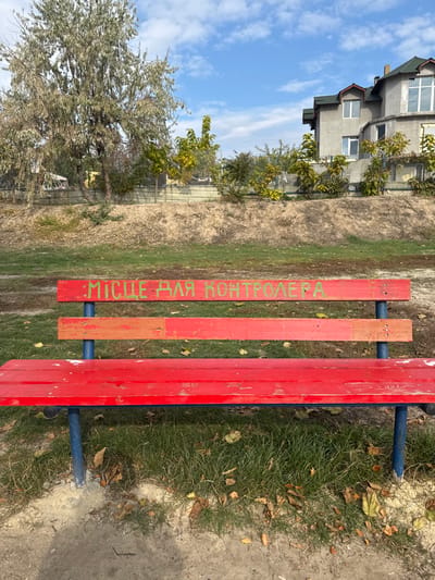 An outdoor scene in Pidhorodnie, Ukraine, is depicted under a partly cloudy blue sky. The central subject is a weathered red wooden park bench, featuring red-painted slats for the seat and backrest, supported by blue metal legs. The top backrest slat displays green Cyrillic text: "МІСЦЕ ДЛЯ КОНТРОЛЕРА," translating to "PLACE FOR THE CONTROLLER." The bench's red paint exhibits wear, with areas of fading and chipping. The ground immediately surrounding the bench consists of patchy green grass, exposed earth, and scattered dry fallen leaves. In the mid-ground, a raised dirt embankment topped by a metal fence is visible, partially obscured by sparse bushes and young trees. The background on the left features large deciduous trees with minimal foliage, while the right background shows a multi-story residential building with light grey stucco walls, a dark green pitched roof, and multiple windows. No individuals or active interactions are present in the image.