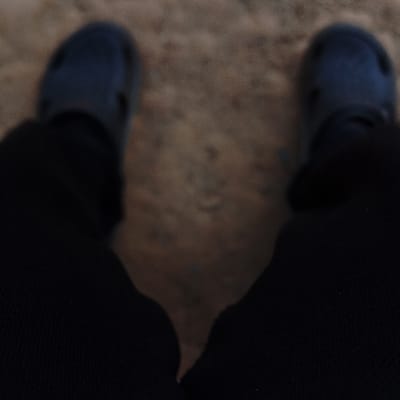 A blurred, first-person, downward-facing perspective captures an individual's lower extremities. Two feet are visible, each wearing a dark blue, closed-toe clog (identifiable as Crocs-style footwear). The feet rest on a light brown, granular surface consistent with dirt or sand. Dark, possibly black, fabric trousers cover the legs, obscuring the ankles and calves. No other individuals or specific objects are discernibly clear due to the heavy blur. While the location is stated as Sámara, Costa Rica, the image lacks specific visual cues to corroborate this geographical detail.