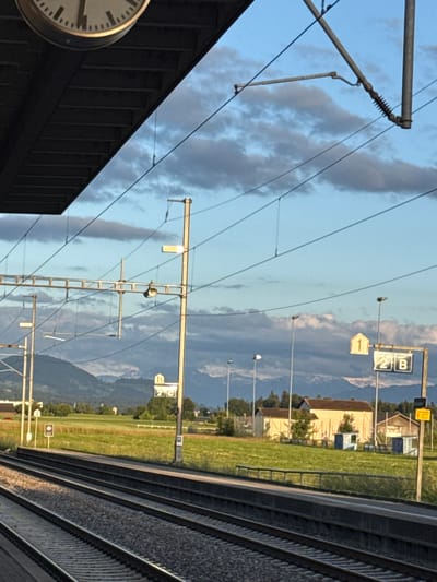 This wide-angle image, rotated 90 degrees, depicts a railway track area, likely a station in Fehraltorf, Switzerland. The scene is dominated by railway infrastructure set against a backdrop of natural landscapes.
On the left side, the edge of a dark, modern building, possibly a station building or platform shelter, is visible. A large, round station clock with Roman numerals, partially visible, is affixed to this building, which features a ribbed or slatted facade. Stretching across the mid-ground are numerous overhead power lines (catenary) supported by tall metal poles and intricate structures, including insulators. A speaker or horn is attached to one of these catenary poles.
Various informational signs are mounted on the poles. One prominent yellow sign shows an upward arrow and the number "1". Another rectangular sign displays "2 B", likely indicating a platform or track designation. Further in the background, a smaller sign shows "15" and possibly "2 C".
To the right, several parallel railway tracks are visible, laid on gravel ballast, receding into the distance. Beyond the tracks and infrastructure, the background reveals vast green fields dotted with a few scattered buildings. Majestic mountains rise in the far distance, under a sky that is partly blue with scattered, darker clouds. The lighting suggests it is late afternoon or early evening. No people or trains are visible, but the presence of the tracks and station elements indicates an active railway environment.