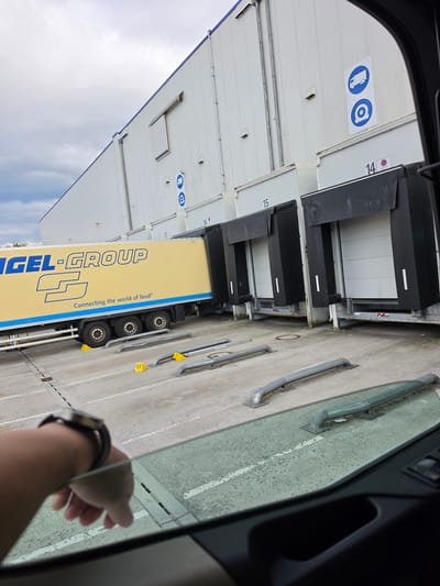 The image shows a yellow semi-trailer truck from "ENGEL-GROUP" parked at a loading dock of a large, white warehouse building. The truck has the slogan "Connecting the world of food®" printed on its side. Several loading bay doors are visible, some with black shelters. Signs are mounted on the warehouse wall, including blue circular signs depicting a truck and a wheel. The loading area is paved with concrete and marked with lines. There are yellow wheel chocks placed in front of the truck's wheels. The view is from inside a vehicle, looking out through the windshield. A person's arm wearing a watch is visible resting on the window frame. The sky is overcast. This scene is likely at a logistics center or distribution facility. The question states the location is Flörsheim, Germany, implying this is a view of a facility within or near that city.
