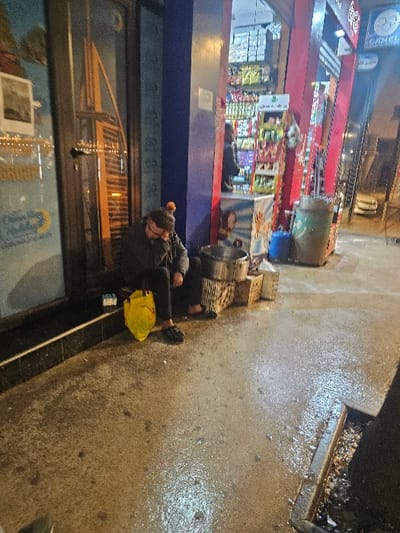 A person is crouched beside a storefront with a yellow bag nearby on a wet city sidewalk. Adjacent to them are metal bins and various stacked items. The storefront, behind the person, displays a depiction of a bridge in its window. A store selling goods is visible, with various products on display and people inside. The storefront is under a sign. A street drain is located on the right edge of the image.